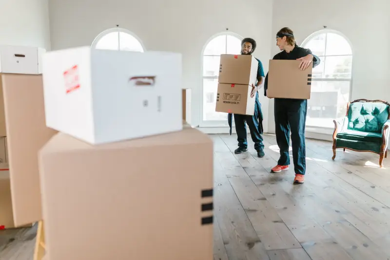 Two movers carrying cardboard boxes through a bright, empty room